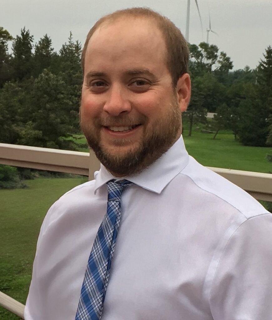 Michael Yerman of My Rural Planner stands outdoors in front of green trees and grass, smiling at the camera—reflecting his passion for community development.